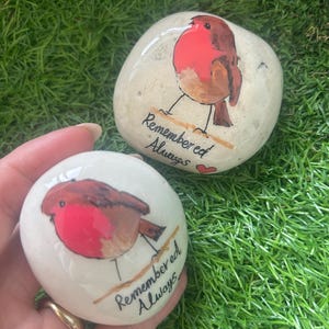 May include: Two smooth, white stones, each featuring a hand-painted robin. The birds have red breasts and brown wings. The text "Remembered Always" is written below each bird. One stone includes a small red heart.