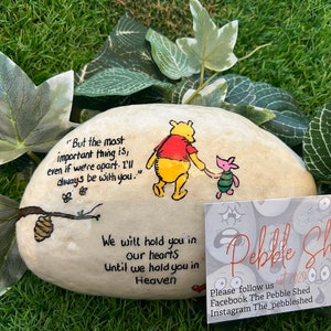 May include: Painted rock featuring Winnie the Pooh and Piglet characters, a quote about friendship, and the text "We will hold you in our hearts Until we hold you in Heaven". The rock is on a green grassy surface with a business card for "Pebble Shed" with social media information.