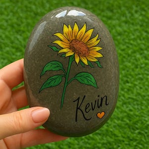 May include: A painted stone with a sunflower design. The sunflower has yellow petals and a brown center, with green leaves and stem. The name "Kevin" is written in cursive with a small orange heart.