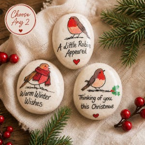 May include: Three smooth, cream-colored oval stones featuring hand-painted illustrations of robins. One stone reads A little Robin Appeared, another Warm Winter Wishes, and the third Thinking of you this Christmas, each accented with small red hearts.