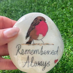 May include: A hand-painted, smooth, white stone with a brown and red robin bird illustration. The words "Remembered Always" are written in cursive, with a small red heart. The stone is held against a background of green grass.