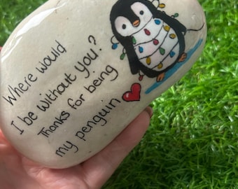 May include: A hand-painted, oval-shaped stone featuring a cartoon penguin adorned with colorful string lights. The stone has the text "Where would I be without you? Thanks for being my penguin" and a red heart.