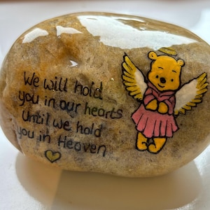 Może przedstawiać: Pomalowany kamień z namalowanym na nim postacią Winnie the Pooh w postaci anioła. Na kamieniu widnieje napis "We will hold you in our hearts until we hold you in Heaven".