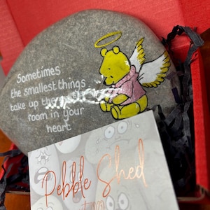 Op de afbeelding: Een grijs geverfde steen met een geverfd Winnie de Poeh-personage erop. Het personage draagt een roze shirt en heeft engelenvleugels. De steen heeft de tekst "Sometimes the smallest things take up the most room in your heart" erop geschilderd. De steen zit in een klein doosje met een visitekaartje waarop "Pebble Shed est 2020" staat en social media-informatie.