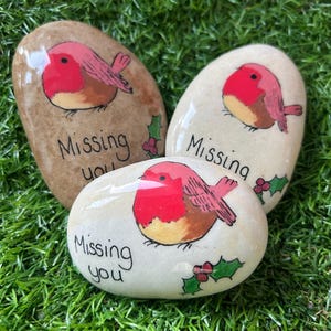 May include: Three painted stones featuring robin bird illustrations and the words "Missing you". The robins have red breasts and brown wings. Holly sprigs are also painted on the stones, set against a white or brown background.