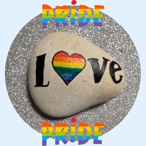May include: A white painted rock with the word "LOVE" written in black paint. A rainbow heart is painted in the center of the rock. The words "PRIDE" are written in rainbow letters above and below the rock.