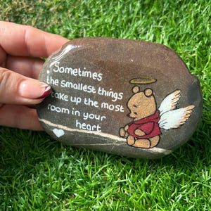 以下が含まれることがあります： 「Sometimes the smallest things take up the most room in your heart」の文字が書かれた、絵が描かれた石。天使の羽と光輪を持つ漫画のクマが描かれています。石は茶色で、緑の草の上に置かれています。