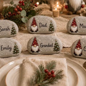 May include: Five smooth, oval-shaped grey stones used as place settings, each featuring a hand-painted gnome in a red hat, a green Christmas tree, and a personalised name in black script. The names include Sarah, Dad, Mum, Emily, Grandad, and Oliver.