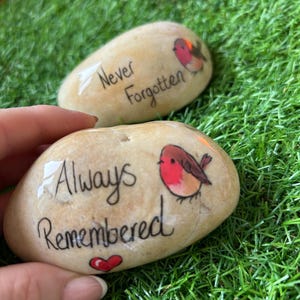 Puede incluir: Dos piedras lisas de color beige con texto manuscrito e ilustraciones. Una piedra dice "Never Forgotten" con un pequeño gráfico de pájaro. La otra dice "Always Remembered" con un corazón rojo y un gráfico de pájaro. Las piedras están sobre hierba verde.