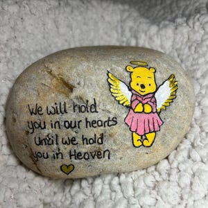 Może przedstawiać: Pomalowany kamień z postacią Winnie the Pooh namalowaną jako anioł z skrzydłami. Na kamieniu widnieje napis "We will hold you in our hearts until we hold you in Heaven".