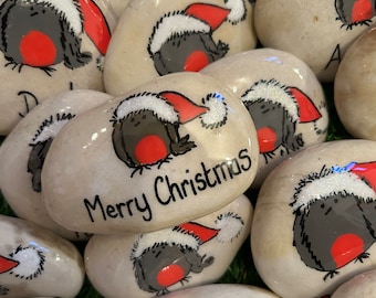 Può includere: Una collezione di pietre dipinte con un pettirosso che indossa un cappello di Babbo Natale. Le pietre sono decorate con glitter e le parole "Buon Natale".