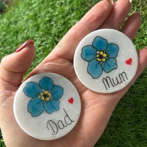 May include: Two circular white stones, each featuring a hand-painted blue forget-me-not flower and a red heart. One stone has the word "Dad" and the other "Mum" in black script. The stones are held in a hand against a backdrop of green grass.