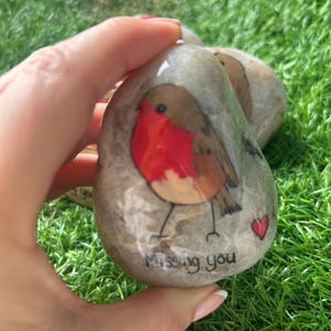 May include: Hand-painted stone featuring a robin design. The bird has a red breast and brown wings, set against a gray stone. The text "Missing you" and a red heart are painted on the stone. The stone is held against a backdrop of green grass.