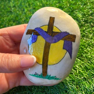 May include: A painted rock with a brown cross and a purple cloth draped over it. The cross is in front of a yellow circle with a white halo. The rock is painted with green grass at the bottom.