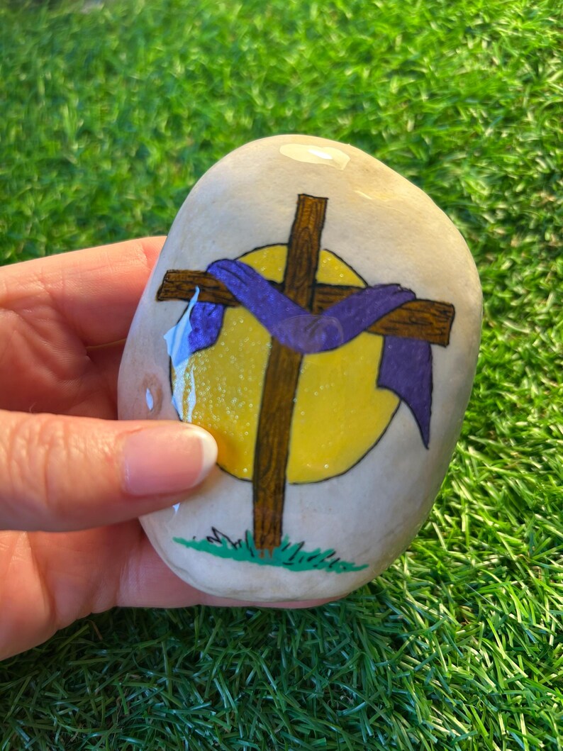 May include: A painted rock with a brown cross and a purple cloth draped over it. The cross is in front of a yellow circle with a white halo. The rock is painted with green grass at the bottom.