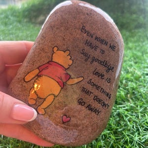 Op de afbeelding: Een handbeschilderde, bruine steen met een cartoonbeer in een rood shirt. De steen heeft de tekst "Even when we have to say goodbye love is something that doesn't go away" en een klein rood hart.