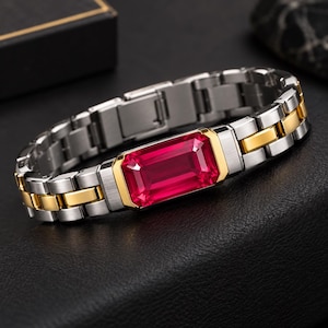 Könnte beinhalten: Ein Gliederarmband mit einem zentralen, rechteckigen roten Edelstein in einer goldfarbenen Fassung. Das Armband besteht aus abwechselnden Gliedern aus gebürstetem silberfarbenem und poliertem goldfarbenem Metall im Uhren-Design.