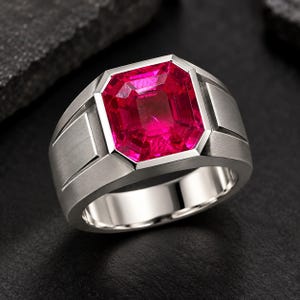 Peut inclure: Une bague argentée ornée d'une grande pierre précieuse rectangulaire à facettes d'un rose vif, montée dans un serti clos. L'anneau présente une finition brossée mate avec des lignes géométriques nettes et un intérieur poli, offrant un aspect structuré.