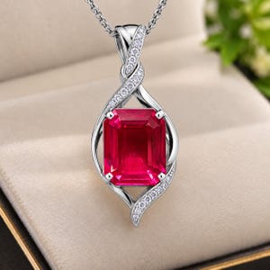 May include: A silver-toned pendant necklace featuring a large, rectangular, deep red gemstone set in a twisted metal frame. The frame is accented with small, clear, sparkling stones along the curved edges, suspended from a delicate silver chain.