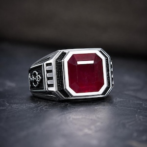 May include: A silver-toned ring featuring a large, rectangular, deep red gemstone set in the centre. The band is crafted with a textured black finish on the sides, accented by intricate silver metalwork and a geometric, stepped-edge design.