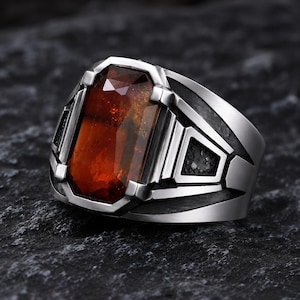 May include: A silver-toned metal ring featuring a large, rectangular, faceted amber-colored stone set in the center. The band has a complex, geometric design with recessed black detailing and angular metal accents surrounding the central gemstone.