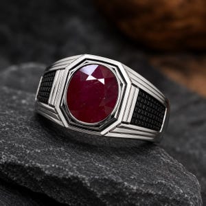 May include: A silver-toned ring featuring a large, oval-cut, deep red gemstone set in the centre. The band has a geometric, stepped design with black, grid-textured panels on the sides, creating a structured and layered appearance.