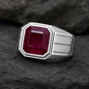 May include: A silver-coloured ring featuring a large, square-cut deep red gemstone set in a stepped bezel. The band has a brushed metal finish with three horizontal grooved lines on each side, creating a structured and geometric appearance.