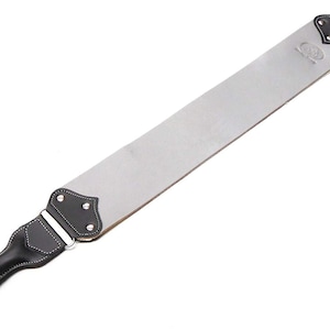 Pode incluir: Uma tira de couro cinza com uma alça de couro preto e um laço de metal prateado. A tira é usada para afiar navalhas de barbear.