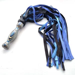 May include: A handcrafted flogger with a wooden handle featuring blue and white swirls. The flogger's falls are a mix of black, light blue, and dark blue leather strips, creating a striking contrast. The handle is wrapped in black leather.
