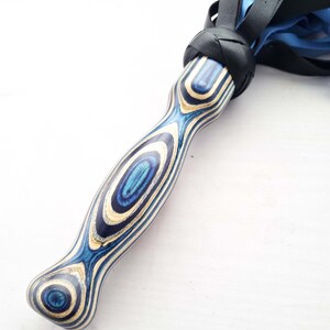 May include: A handcrafted flogger with a blue and white wooden handle. The handle has a unique grain pattern and is connected to black leather and blue falls. The flogger is designed for impact and sensory play.