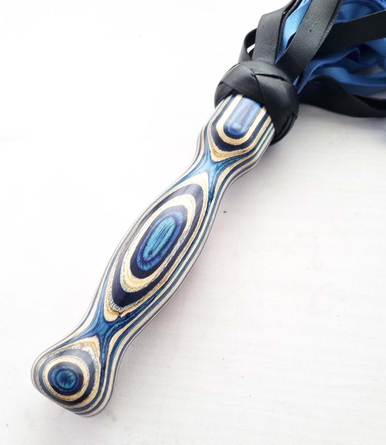 May include: A handcrafted flogger with a blue and white wooden handle. The handle has a unique grain pattern and is connected to black leather and blue falls. The flogger is designed for impact and sensory play.