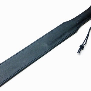May include: A black leather paddle with a rounded handle and a rectangular striking surface. The edges are stitched with black thread. A black cord is attached to the handle.
