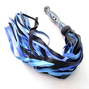 May include: A flogger with a wooden handle featuring a blue and white swirl pattern. The flogger's fall is composed of black and shades of blue leather strips, knotted at the base of the handle.
