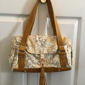 May include: A mustard yellow and white floral print handbag with cork accents and a tassel. The bag has a top flap closure and two straps.