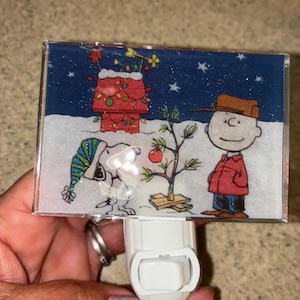 Peut inclure: Veilleuse rectangulaire avec une scène festive mettant en vedette Snoopy, Charlie Brown et un sapin de Noël. Snoopy porte un chapeau bleu et vert, et Charlie Brown est en veste rouge. Le fond est un ciel étoilé avec un sol enneigé.