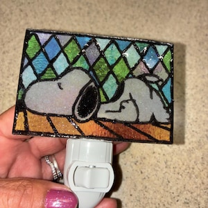 May include: A rectangular night light featuring a stained glass style illustration of the cartoon character Snoopy sleeping on a brown surface against a background of blue and green diamond shapes with a white plastic plug-in base.