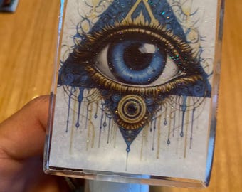 Puede incluir: Una luz nocturna con una cubierta de plástico transparente que presenta un ojo azul dentro de un triángulo dorado sobre un fondo azul y dorado. El ojo está rodeado de detalles dorados y azules. La luz nocturna está sobre una superficie de madera.