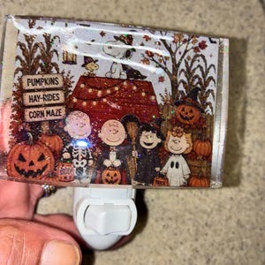 May include: A rectangular night light featuring a Peanuts Halloween scene with Snoopy, Charlie Brown, and friends in costumes. The design includes pumpkins, a sign reading PUMPKINS, HAY-RIDES, CORN MAZE, and a red doghouse, all covered in a holographic glitter finish.