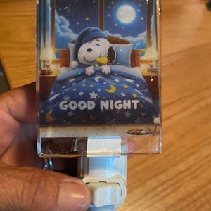 Puede incluir: Una luz nocturna blanca con un inserto de plástico transparente que presenta una imagen de dibujos animados de Snoopy en la cama con el texto "Buenas noches".