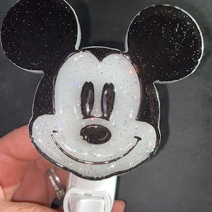 Puede incluir: Un enchufe de luz nocturna blanco con un diseño de Mickey Mouse de purpurina negro y blanco.