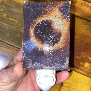 May include: A nightlight with a clear resin cover featuring a glittery image of a solar eclipse. The nightlight is white and has a plug-in design.