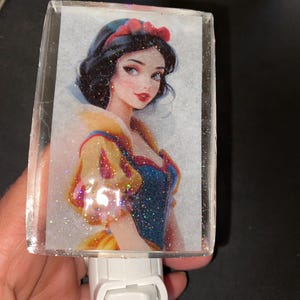 May include: A white night light with a clear plastic cover featuring a glitter-covered image of Snow White. The image shows Snow White with a red headband and a yellow and blue dress. The night light has a white on/off switch.