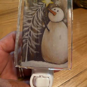 May include: A white snowman with a carrot nose and a yellow star on top of his head. The snowman is standing in front of a white pine tree with a brown wooden background. The image is on a clear glass nightlight.