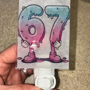 May include: A night light featuring the numbers 67, designed as cartoon characters with arms, legs, and shoes. The numbers are a gradient of pink and blue with a dripping effect, and are set against a glittery background.