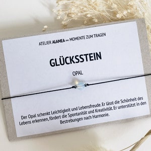 Könnte beinhalten: Ein schwarzes Armband aus Schnur mit einem hellblauen Opal-Edelstein. Das Armband liegt auf einer Karte mit dem Text "Atelier Alamea - Momente zum Tragen" und "Glücksstein Opal". Die Karte enthält auch eine Beschreibung der Bedeutung des Opals.