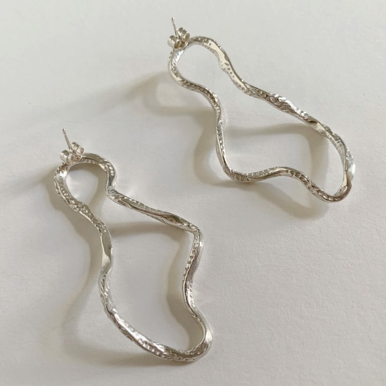 May include: A pair of silver earrings with a wavy, organic shape. The earrings are textured and have a slightly hammered finish.