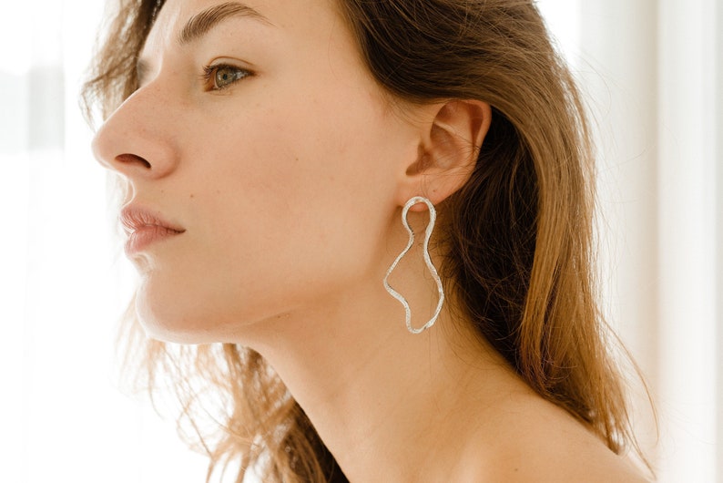 May include: A woman with long brown hair wearing silver abstract earrings. The earrings are shaped like a wavy, organic form.