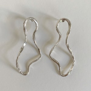 May include: A pair of silver earrings with a wavy, abstract design. The earrings are made of a textured metal and have a unique, organic shape.