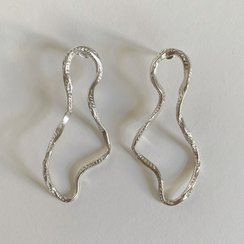 May include: A pair of silver earrings with a wavy, abstract design. The earrings are made of a textured metal and have a unique, organic shape.