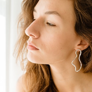 May include: A close-up of a woman's face with long brown hair. She is wearing a silver, abstract-shaped earring.